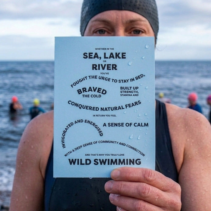 A person in a dark swim cap and wetsuit holds up a light blue printed card with black text reading 'WILD SWIMMING' and 'SEA, LAKE, RIVER' in front of an outdoor water scene at the beach.