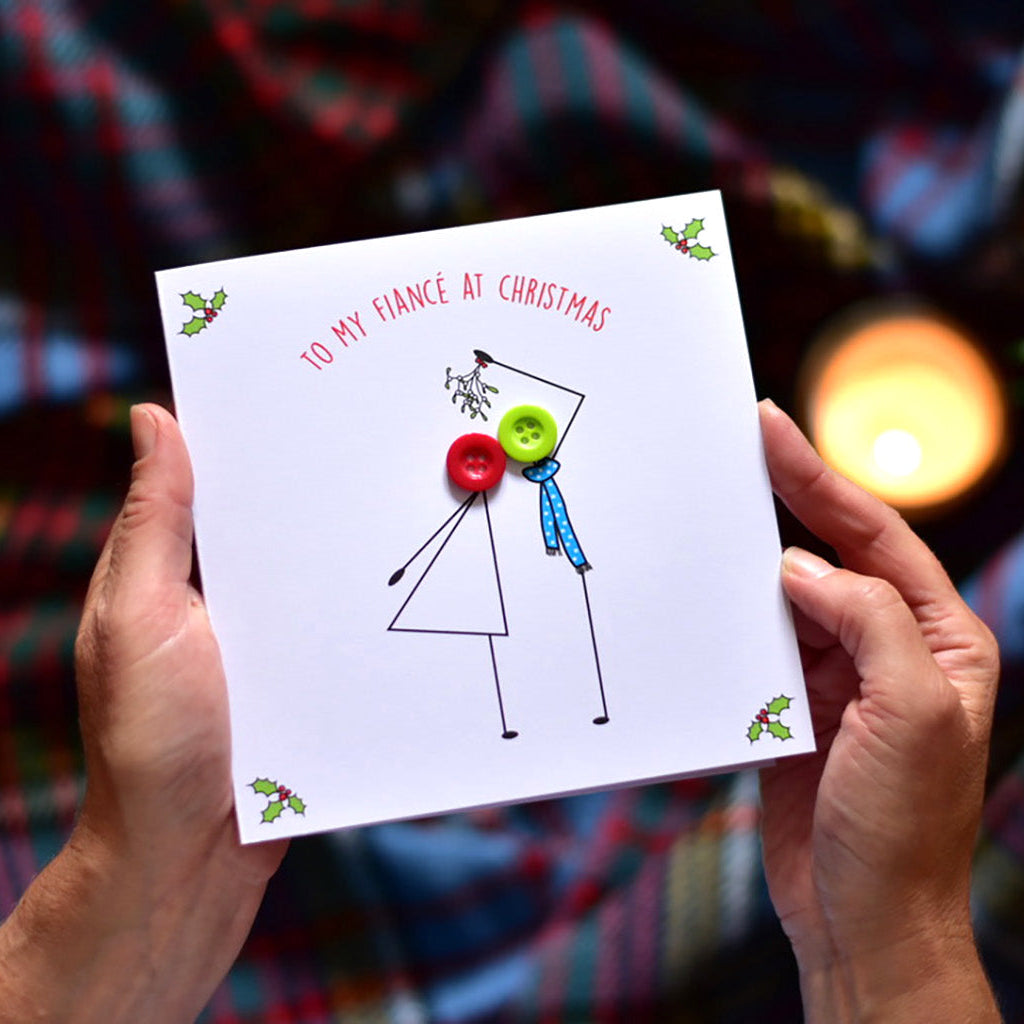 A person holds a white Christmas greeting card with a minimalist stick figure couple illustration and the text 'TO MY FIANCE AT CHRISTMAS' in red. The background shows a cozy, blurred holiday scene with a lit candle and a red plaid blanket.