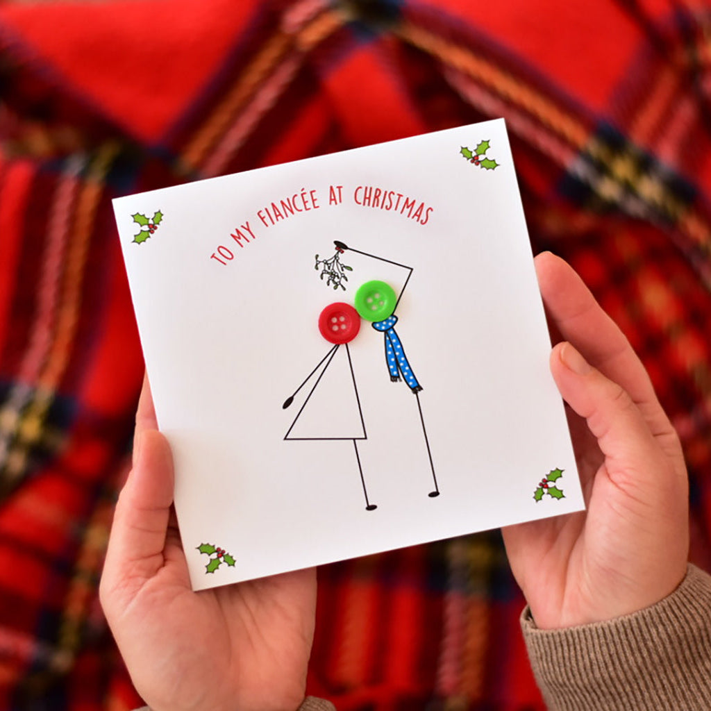 A person holds a white Christmas greeting card that reads 'TO MY FIANCÉE AT CHRISTMAS' with a simple stick figure and holiday accents. The background is a blurred red and black plaid fabric.